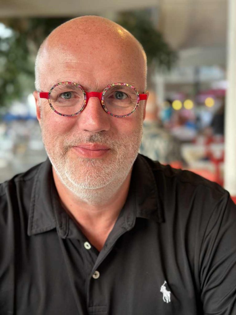 Jens Hövelmann, ein weißer Mann mit bunter Brille und fast keinen Haaren, 3-Tage Bart und schwarzen Polo-Shirt